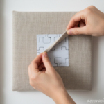 Close-up of hands peeling adhesive backing from a fabric upholstered wall panel, showing the self-ad