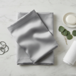 Overhead flat lay of bathroom accessories on white marble: folded shower curtain in grey pearl color