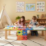 Two children around 4 and 6 years old sitting at a colorful modular puzzle table in a sunny playroom